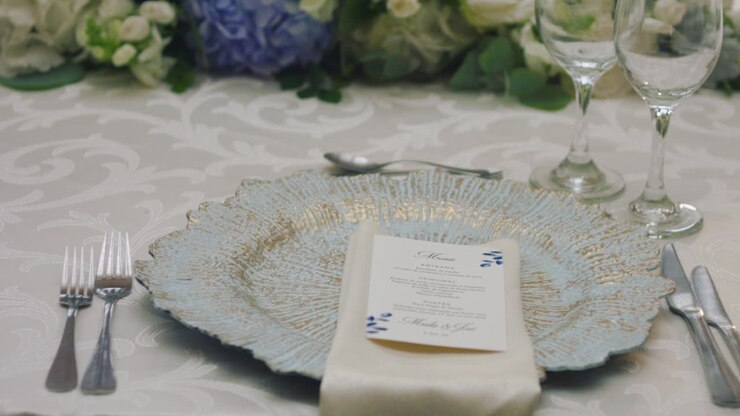 A detailed clip of an elegantly decorated wedding table. It features stemware, a white tablecloth, plates, cutlery, cloth napkins, a delicate floral arrangement, and a personalized invitation.