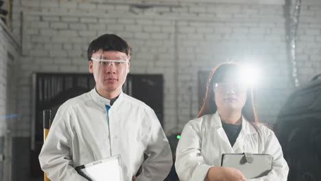 male technician nods with a warm smile while holding a tablet, as his colleague adjusts her safety glasses with a soft smile, white light glows behind her head in an automotive workshop setting