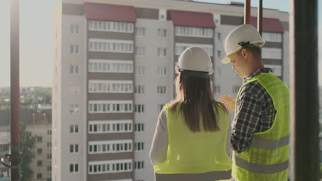 Vista-Posterior,-Joven-Capataz-E-Ingeniero-Revisando-El-Plano-Y-Hablando-Sobre-El-Proyecto-De-Construcción-Con-Compromiso-Con-El-éxito-En-El-Sitio-De-Construcción