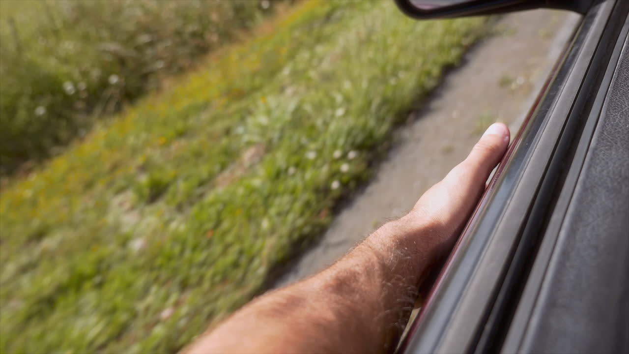 Una mano tamburella sul bordo di un finestrino dell'auto a ritmo, ondeggiando alla musica mentre l'auto si muove. L'auto rossa passa accanto a una sfocatura di vegetazione stradale, catturando un momento spensierato sulla strada.
