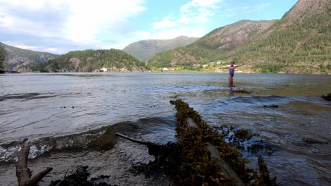 Young-blonde-child-in-swimsuit-playing-in-sea---Static-clip-close-to-water-with-small-waves-rolling-against-camera-and-sunny-mountains-in-background---Western-coast-of-Norway