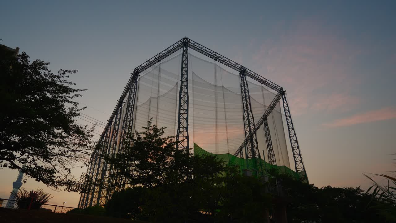 An outdoor golf driving range with a large netting structure, silhouetted against a dusk sky