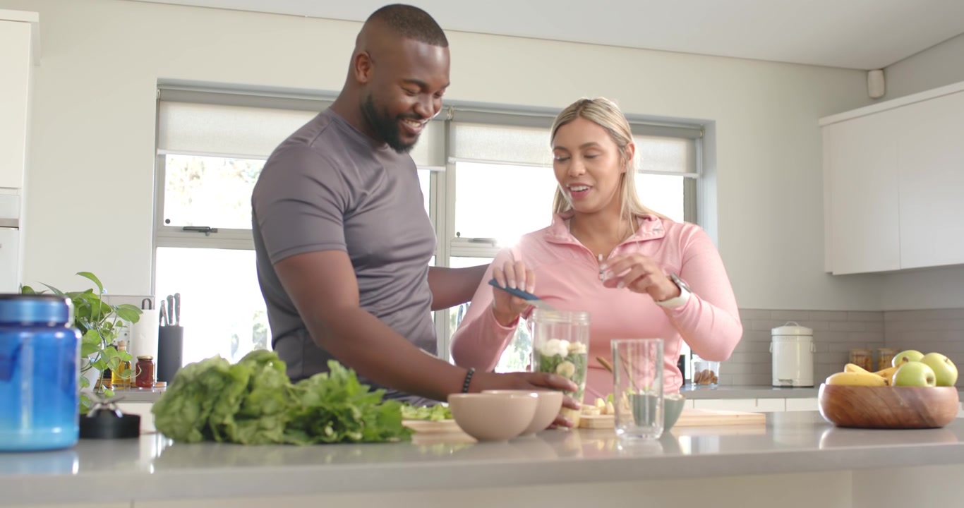 Casal afro-americano, após um carinho, preparando smoothie na ilha da cozinha, adicionando produtos frescos ao liquidificador.