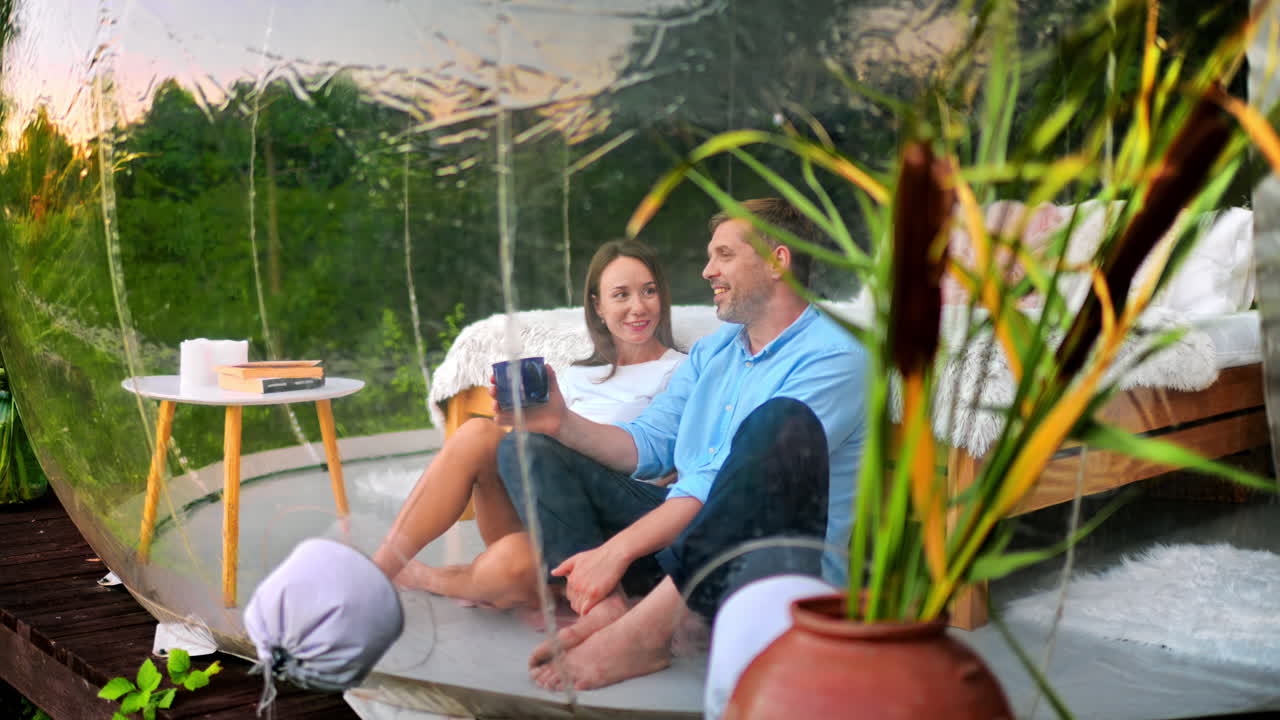A talking couple sitting inside transparent bubble tent at glamping with cups. Lush forest on the background