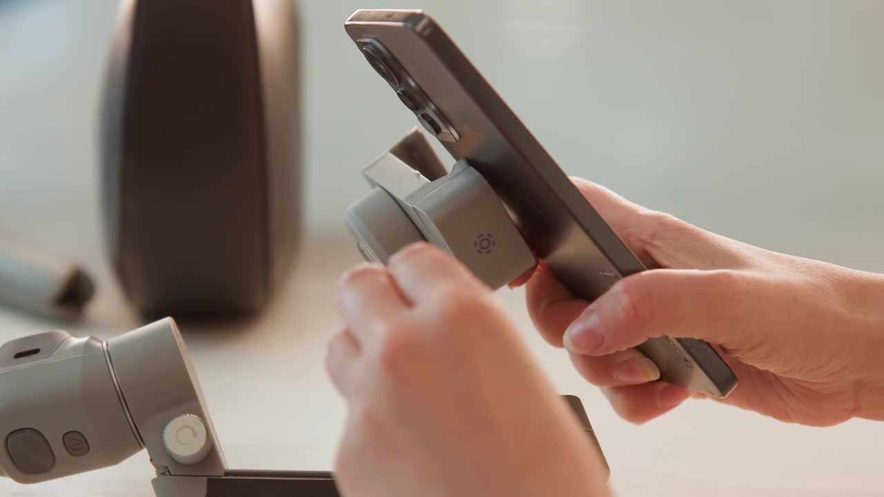 Due mani guidano lo smartphone nel morsetto del gimbal su un bancone, mostrando la pressione delle dita, il movimento del cardine, l'allineamento delle ganasce e una presa sicura durante l'attento montaggio e la rimozione, con mano, smartphone, telefono.