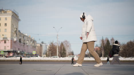 girl in white coat and headset tap dances gracefully on wide tiled plaza framed by tall trees and snow as pedestrians pass on busy city walkway under clear winter sky during daylight