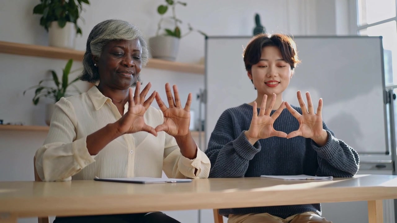 Making two women holding heart shapes after group cue at meeting table, tablet, blouse and sweater