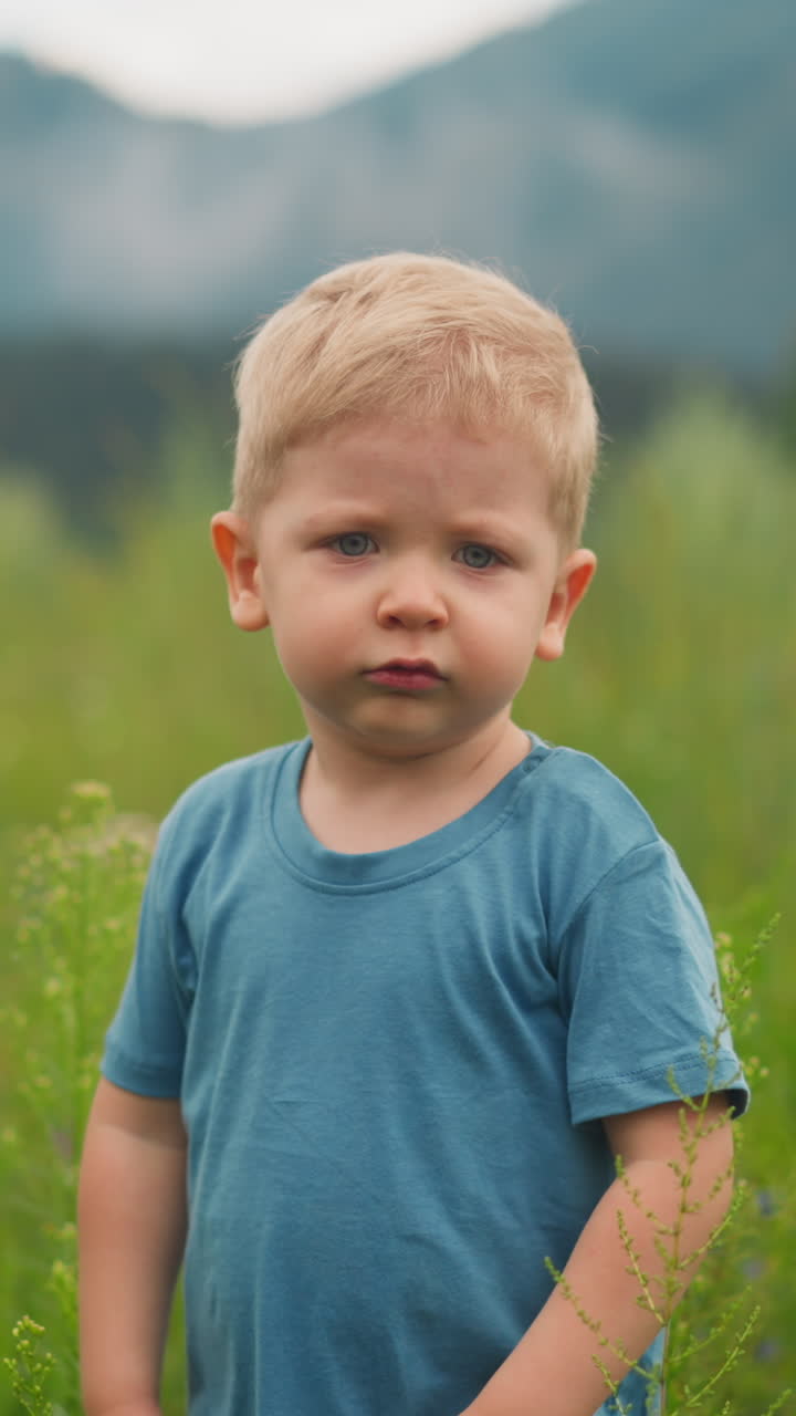 Upset little red-haired boy in blue t-shirt stands among high green grass walking in wild lush field in valley on cloudy day closeup