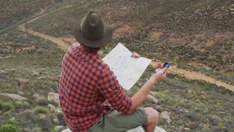 caucasian male survivalist sitting on mountain peak in wilderness, holding map and compass