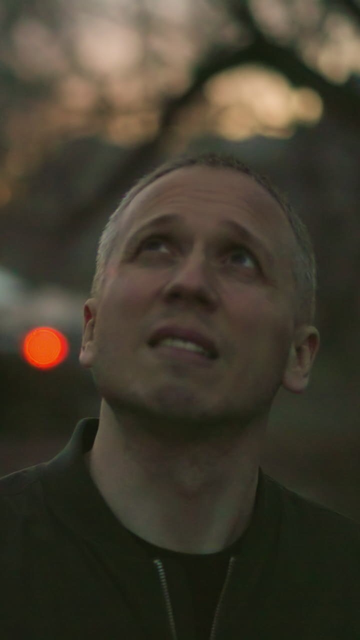A man in a green jacket stands in a dimly lit outdoor setting at dusk, looking upwards with a contemplative expression, smoke is visible around his nose and mouth
