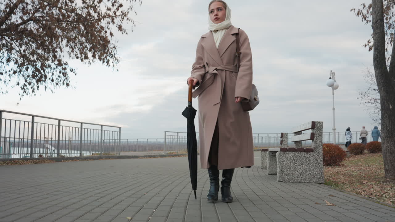 Donna bianca in posa con ombrello, indossa un trench coat beige e stivali, posa da modella editoriale e sguardo calmo, sfondo lungomare, alberi autunnali e cielo nuvoloso, inquadratura ravvicinata del ritratto, elegante borsa in pelle.