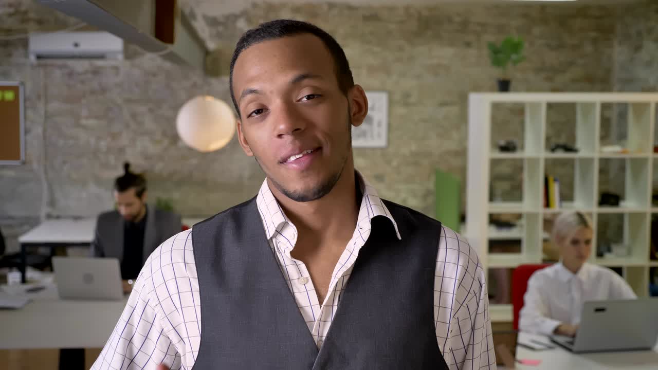 joven empresario africano está de pie y mirando a la cámara en la oficina, mostrando como señal, colegas están trabajando con computadoras portátiles, concepto de trabajo