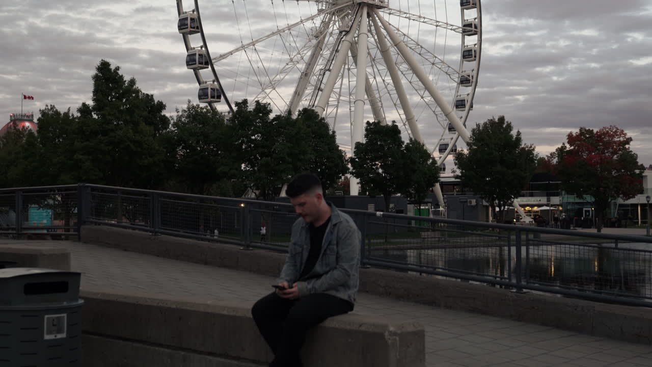 мужчина сидит на каменной ограде и пишет смс на мобильном телефоне - на заднем плане крутится grande roue de montreal во время коронавируса на закате в монреале, канада