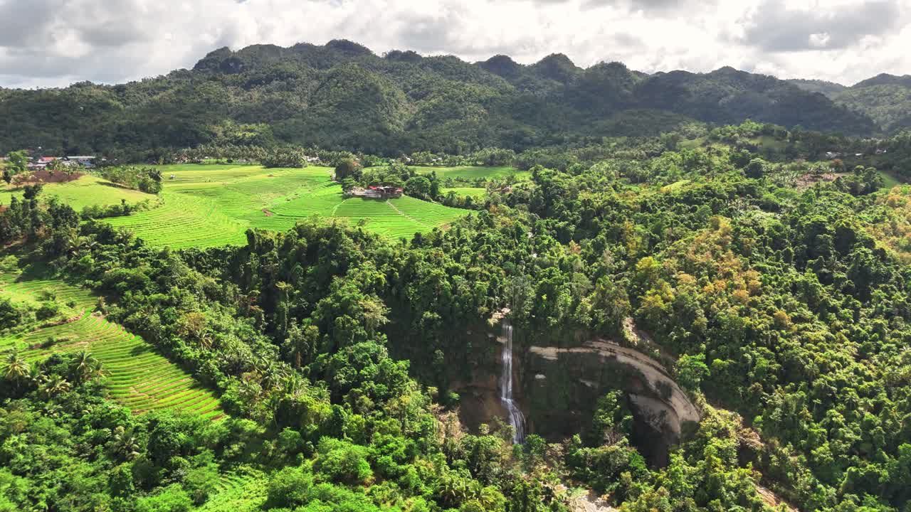 Drone view of Can-umantad Falls cascading beside vibrant Cadapdapan Rice Terraces in Bohol, Philippines, surrounded by dense tropical forest and hills under partly cloudy skies