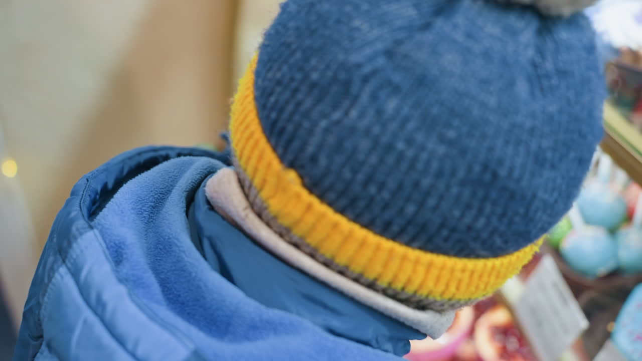 Niño pequeño con gorro de invierno frente a un expositor de pasteles, primer plano de la mano de la mamá guiando a su hijo que presiona la mirada contra el cristal, cupcakes y cake pops coloridos, chaqueta acogedora, luces suaves, momento cándido de anhelo y la madre.