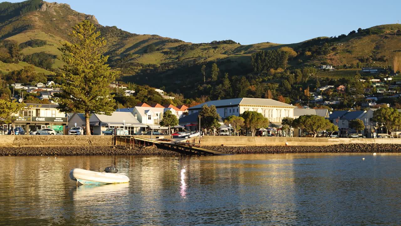 A calm boat drifts on Akaroa's reflective waters during a warm evening, surrounded by picturesque hills and charming seaside shops