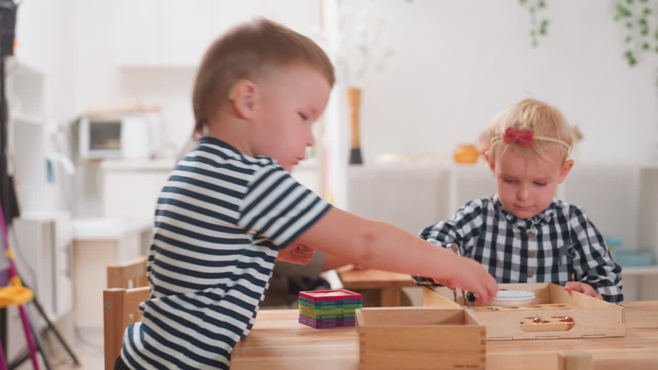 Niño con audífono apila fichas de colores en una mesa de madera durante una sesión preescolar; una profesora le ayuda al fondo mientras una niña rubia con un vestido de cuadros trabaja con una bandeja.