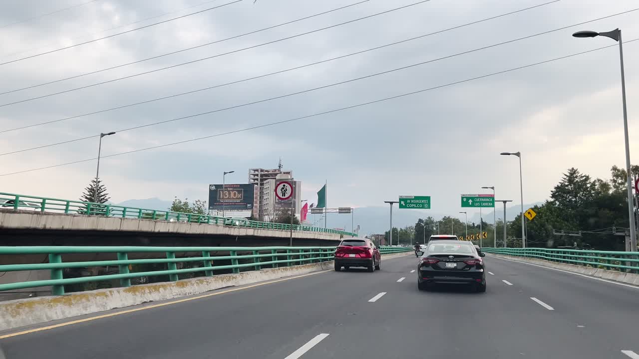 Cars driving on a city highway under a cloudy sky, surrounded by green barriers and urban buildings