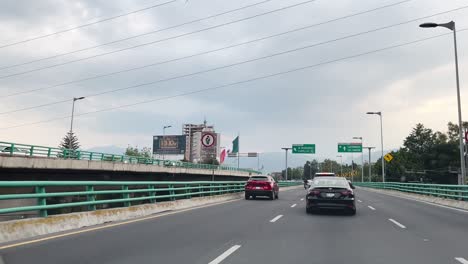 cars driving on a city highway under a cloudy sky, surrounded by green barriers and urban buildings