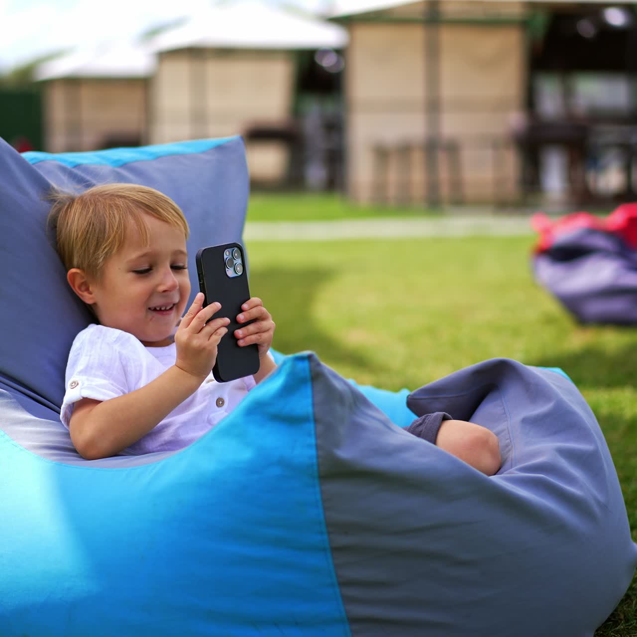 Little boy looking at phone holding it very close to his face. Child in comfortable chair looks aside smiling. Outdoors backdrop in blur