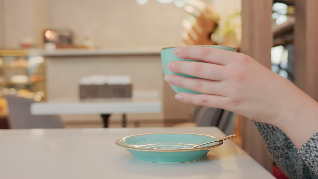 Damsel in cozy cafe gently lifts warm coffee from ceramic cup, takes sip, then places cup back on matching saucer with spoon beside it on table, relaxed ambiance with adjacent table in background