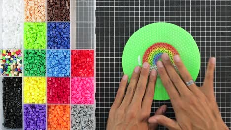 hands crafting a colorful bead rainbow on a green circular pegboard, creativity in progress