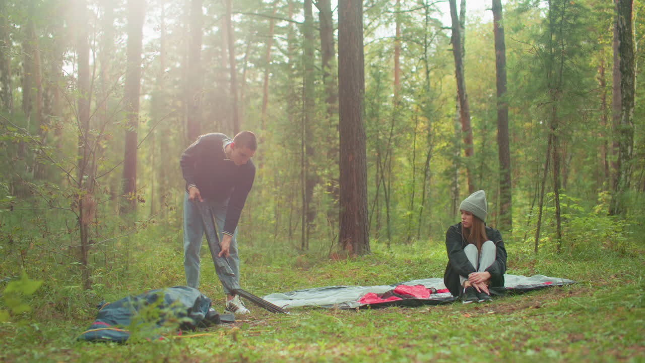 Man squats to unpack and arrange tent poles while woman in gray beanie sits cross-legged on tent fabric watching him in forest, couple setting up for camping surrounded by trees