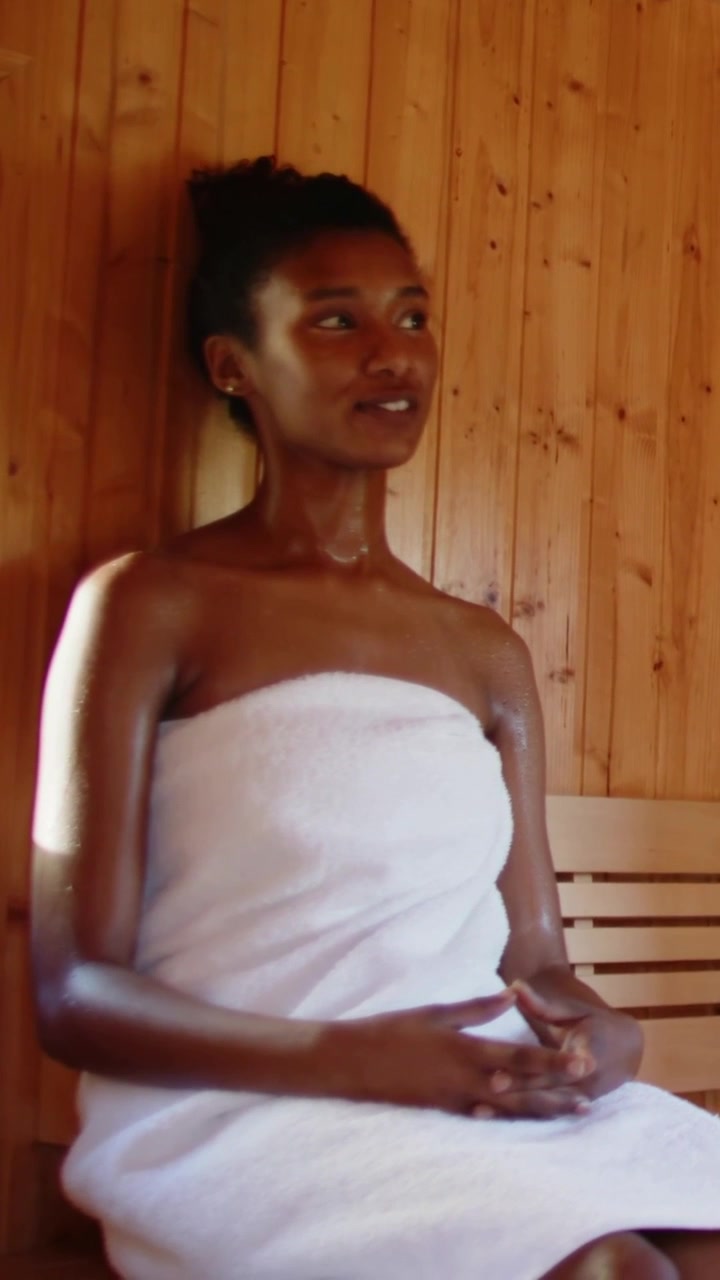 Video verticale: Donna in asciugamano che respira, sposta lo sguardo e si inclina all'indietro per il calore su una panca in sauna.