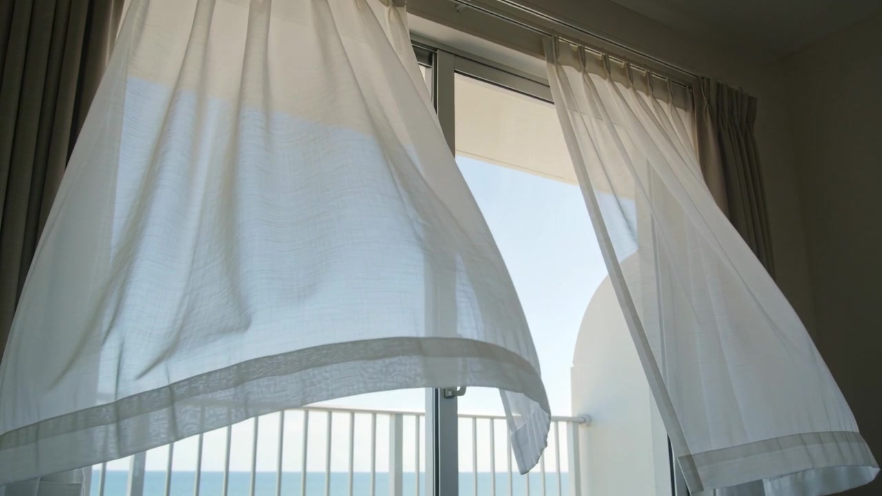 Denizden gelen rüzgar sürgülü kapıdan içeri girdikten sonra dalgalanan şeffaf beyaz perde, balkon korkuluğunu ve denizi gösteriyor.