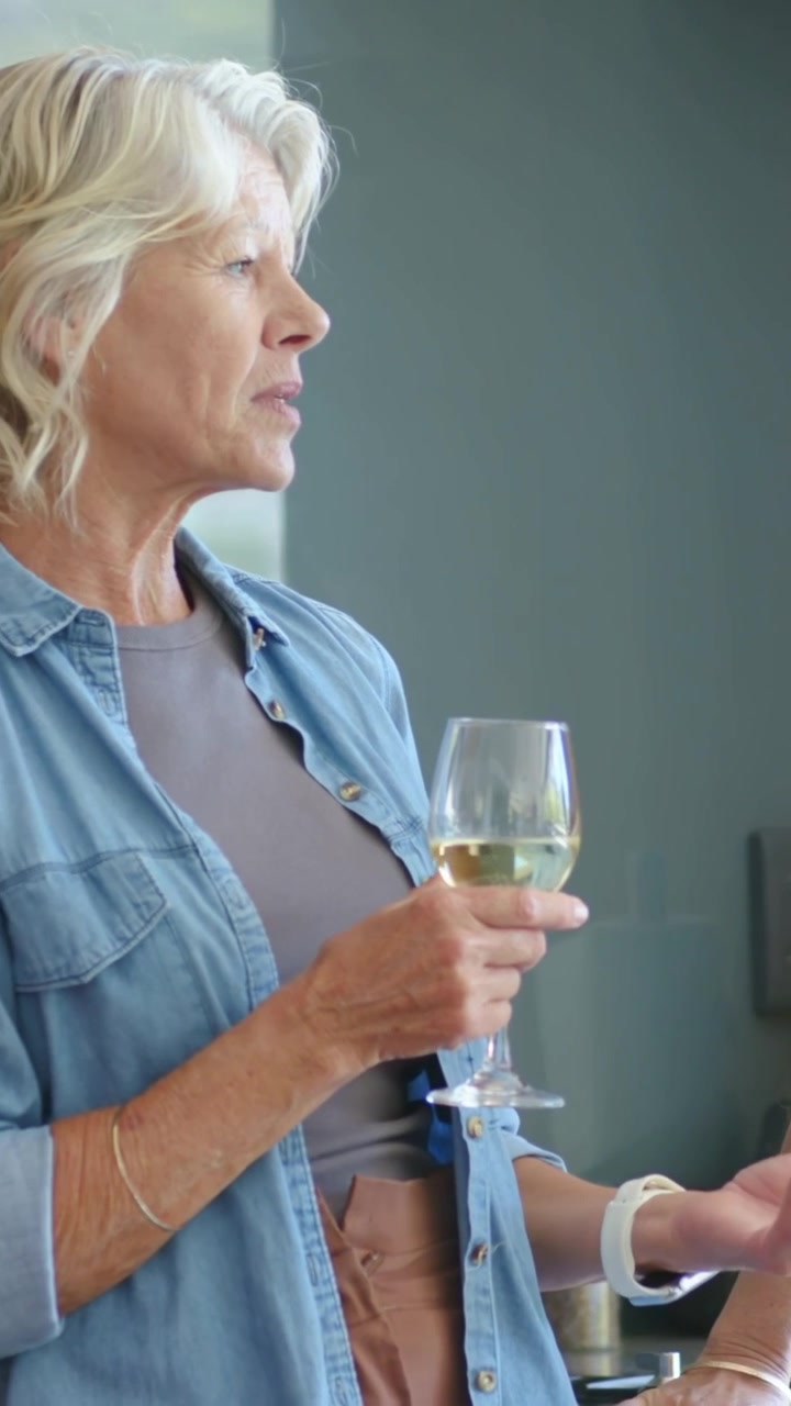 Vertical video: As hand appears woman in denim chatting and gesturing in kitchen with wine glass