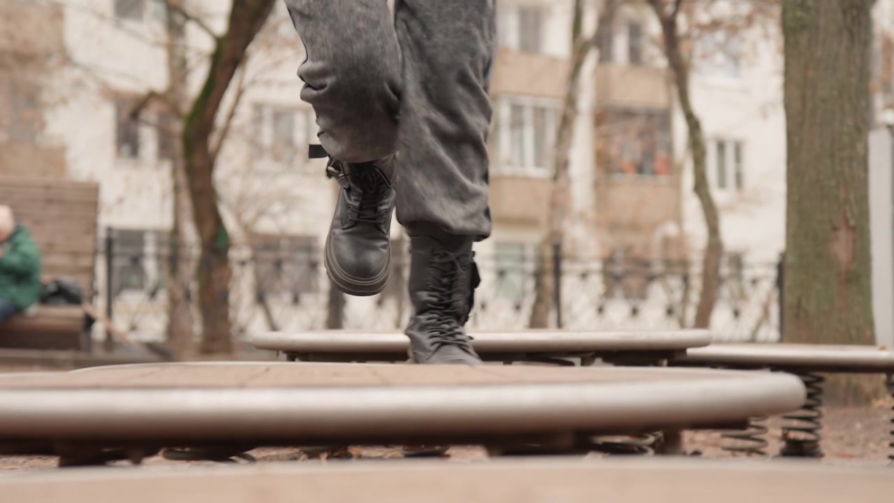 Persona de pie en un parque cerca de una plataforma de resorte, botas pisando un disco circular mientras la plataforma se tambalea ligeramente, luz invernal tenue, árboles desnudos y fachadas de apartamentos distantes enmarcan un momento solitario y contemplativo.