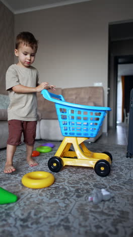 small kid plays with toy shopping cart at home. toddler boy picks the toys from the floor. big suitcase stands beside. vertical video.