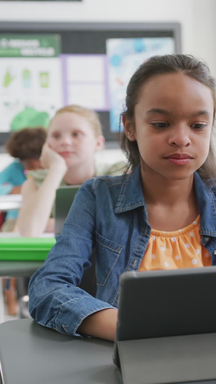 Vertical video of happy bircacial schoolgirl sitting at desk, using tablet in class, copy space