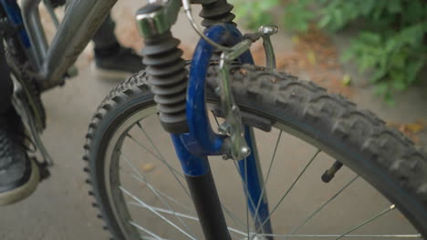 primer plano de alguien probando el freno delantero de una bicicleta en un camino pavimentado, con la banda de rodadura de los neumáticos y la mecánica de los frenos visibles, la vegetación exuberante y las hojas secas esparcidas están presentes en el suelo