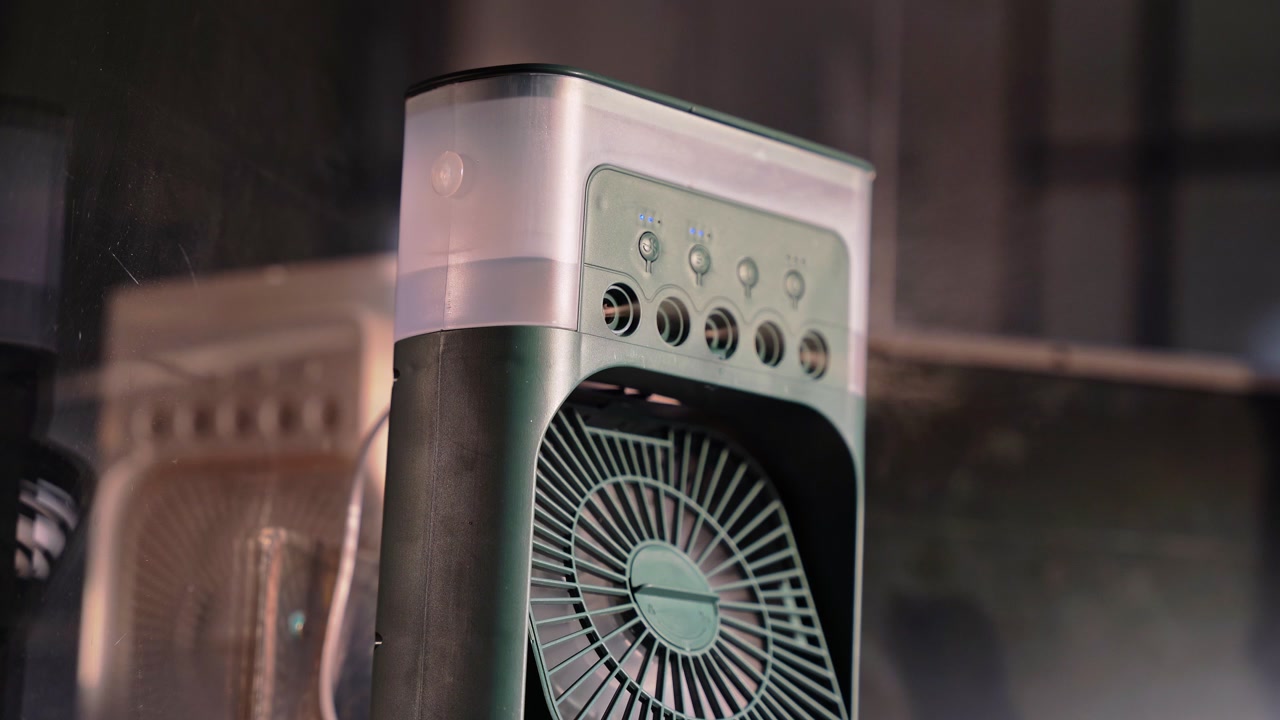 Close up of a portable desktop misting fan in a dim indoor setting. Visible vapor mist spray from a sleek air cooler device with electronic control buttons for personal cooling.