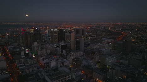 Toma-Panorámica-Elevada-De-Edificios-De-Gran-Altura-En-El-Centro-De-La-Ciudad-Por-La-Noche.-Puerto-En-La-Bahía-Del-Mar-En-El-Fondo.-Ciudad-Del-Cabo,-Sudáfrica