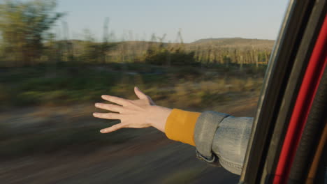 Frau-Im-Auto-Hält-Die-Hand-Aus-Dem-Fenster-Und-Spürt,-Wie-Der-Wind-Durch-Die-Finger-Weht,-Während-Sie-Im-Sommerurlaub-Auf-Dem-Land-Unterwegs-Ist-Und-Bei-Sonnenuntergang-Die-Freiheit-Auf-Der-Straße-Genießt