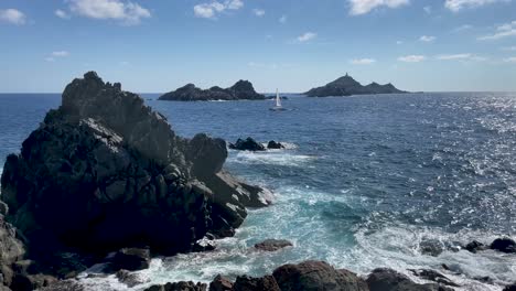 Las-Olas-Del-Mar-Chocan-Contra-La-Costa-Rocosa-De-La-Isla-De-Córcega,-Mientras-Un-Barco-De-Vela-Cruza-El-Mar