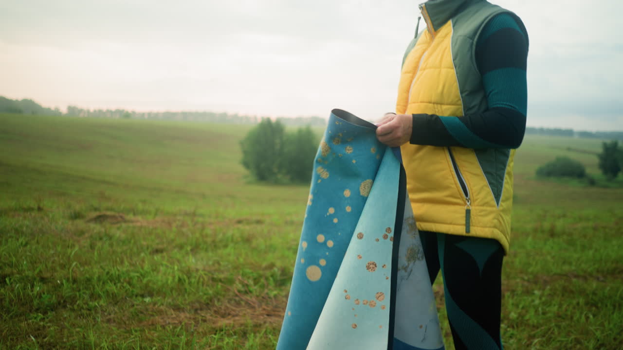 Woman in yellow and green jacket retrieves mat from bag on ground with green coat next to it on grassy field, she picks up mat while looking into the distance with focus, vast field in the background