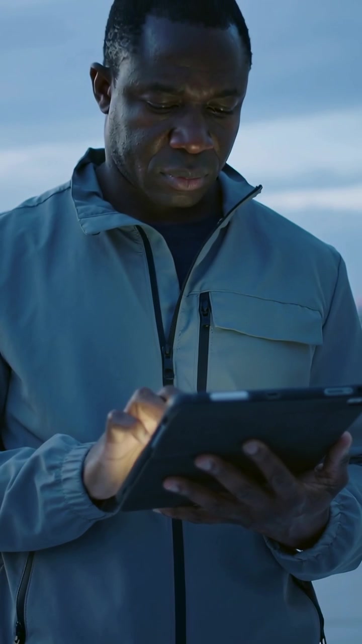 Vertical video: Tapping man in gray zip-up jacket checking messages at shore dusk as tablet glowing