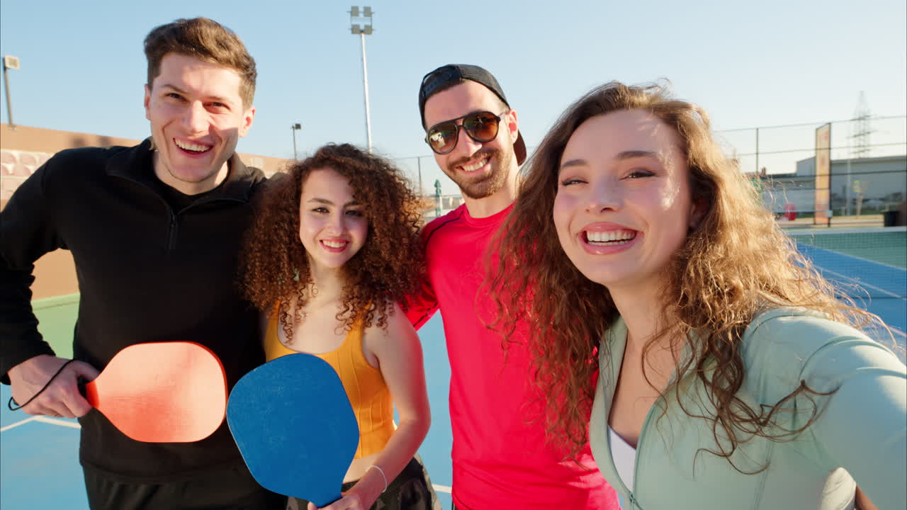 Two men and two women holding pickleball rackets smiling, taking a selfie video on a blue court