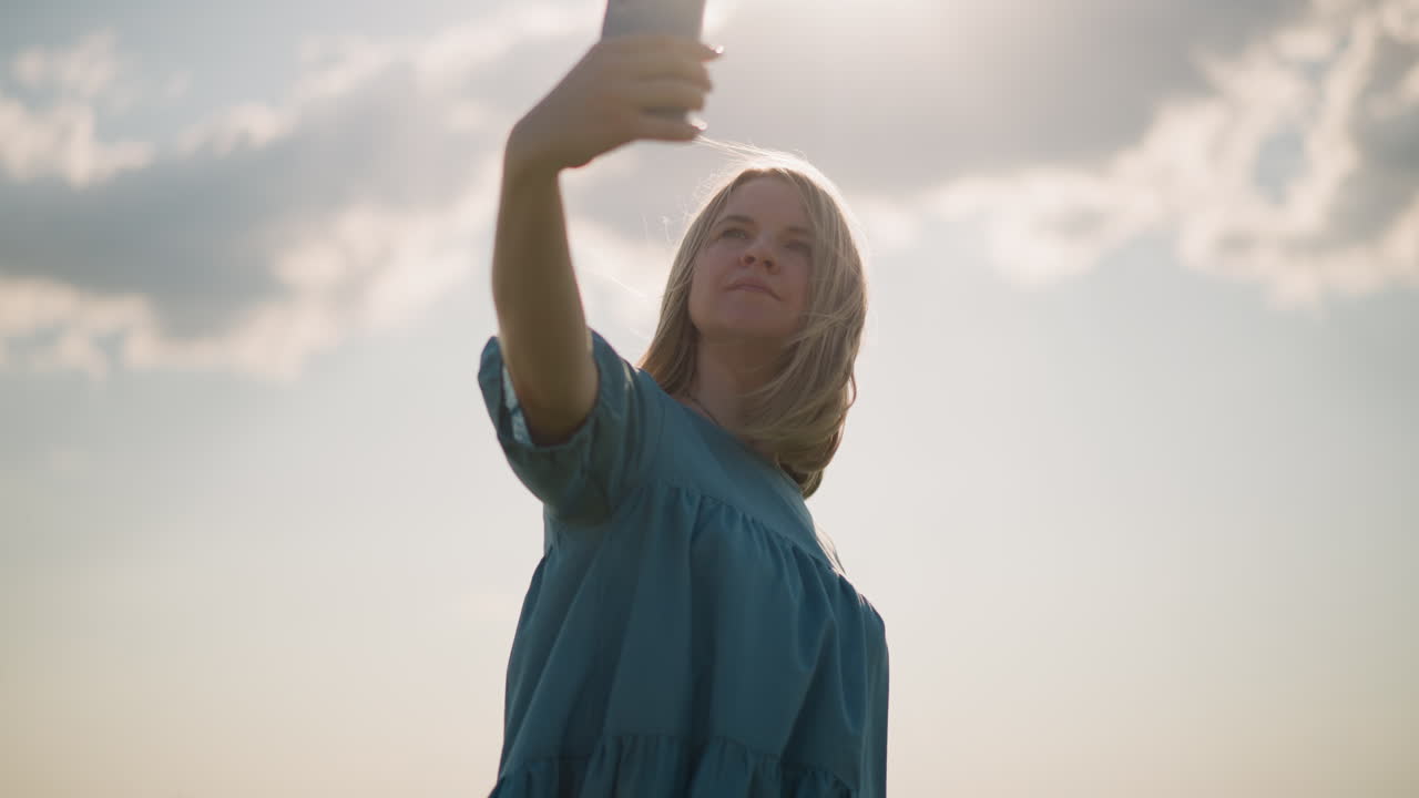 Close-up of a woman wearing a blue dress, holding a smartphone as she films herself outdoors. She gently touches her hair as it blows in the breeze, capturing a moment in the sunlit field