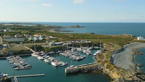 Flight-over-Beaucette-Marina-flying-west-from-out-to-sea,-north-east-coast-of-Guernsey-on-bright-sunny-day-showing-marina-,Fort-Doyle,restaurant,motorhome-campsite-and-views-of-bays-out-to-sea