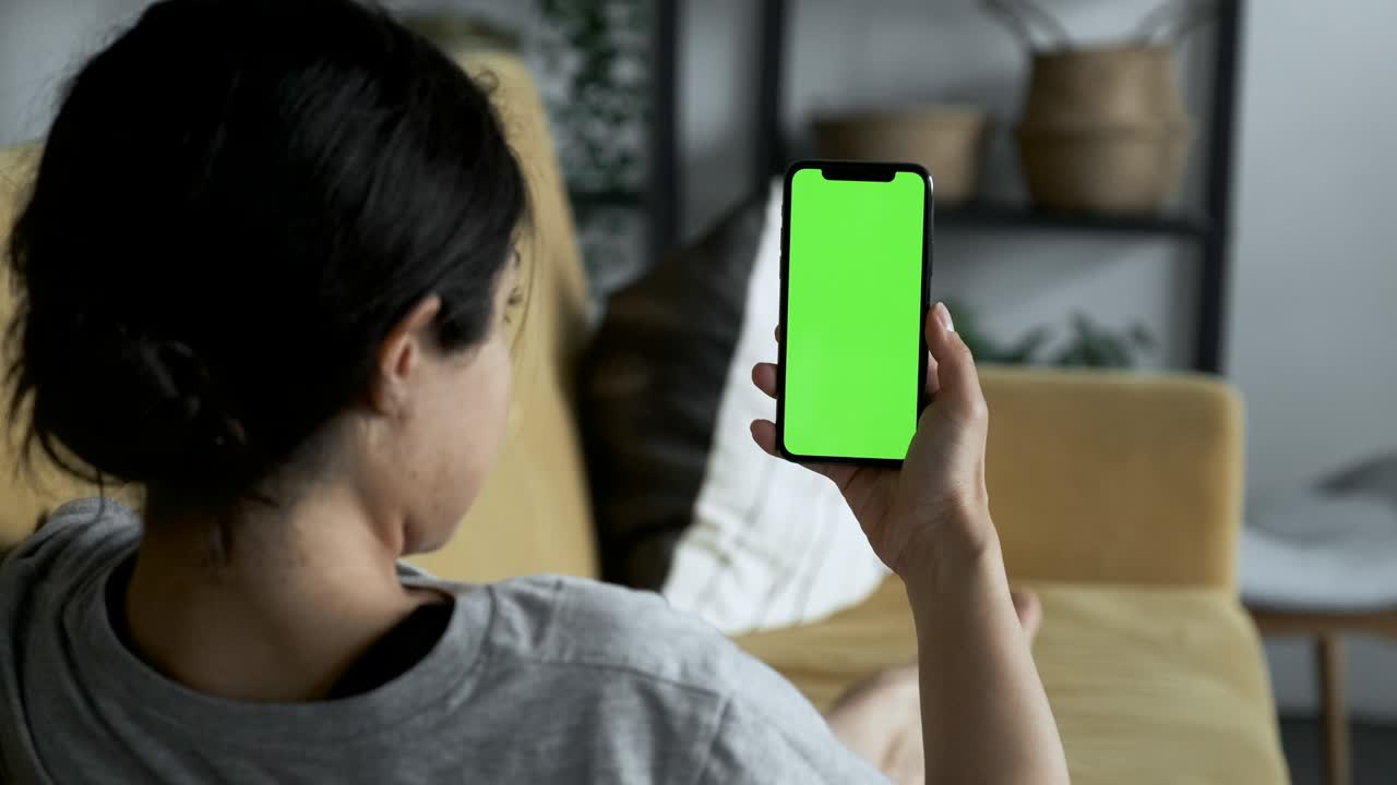 여성의 손은 녹색 화면의 스마트폰을 들고 소셜 네트워크, 영화, 비디오를 보고, 편안한 방의 소파에 누워 있습니다.