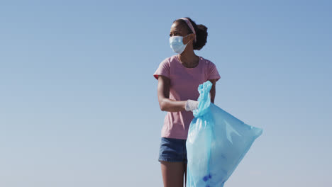 african american woman wearing face mask holding rubbish sack and collecting rubbish from the beach