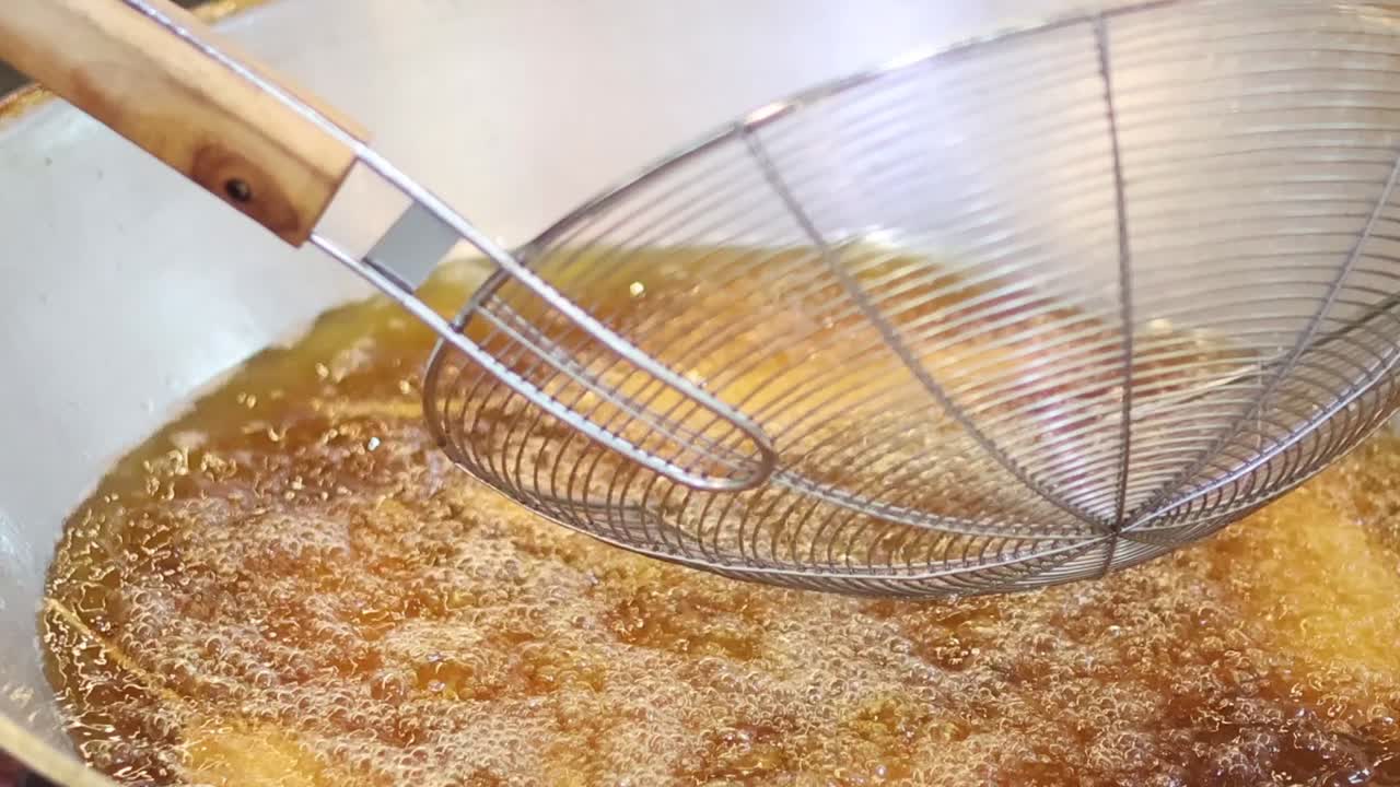 A wire skimmer moves over breaded food deep frying in a large metal wok