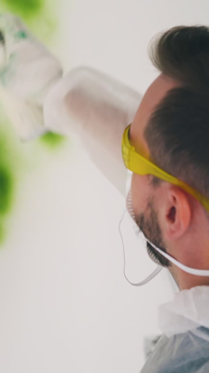 CU: young bearded man in workwear and mask paints white wall using bottle of green spray in repairing room close back view