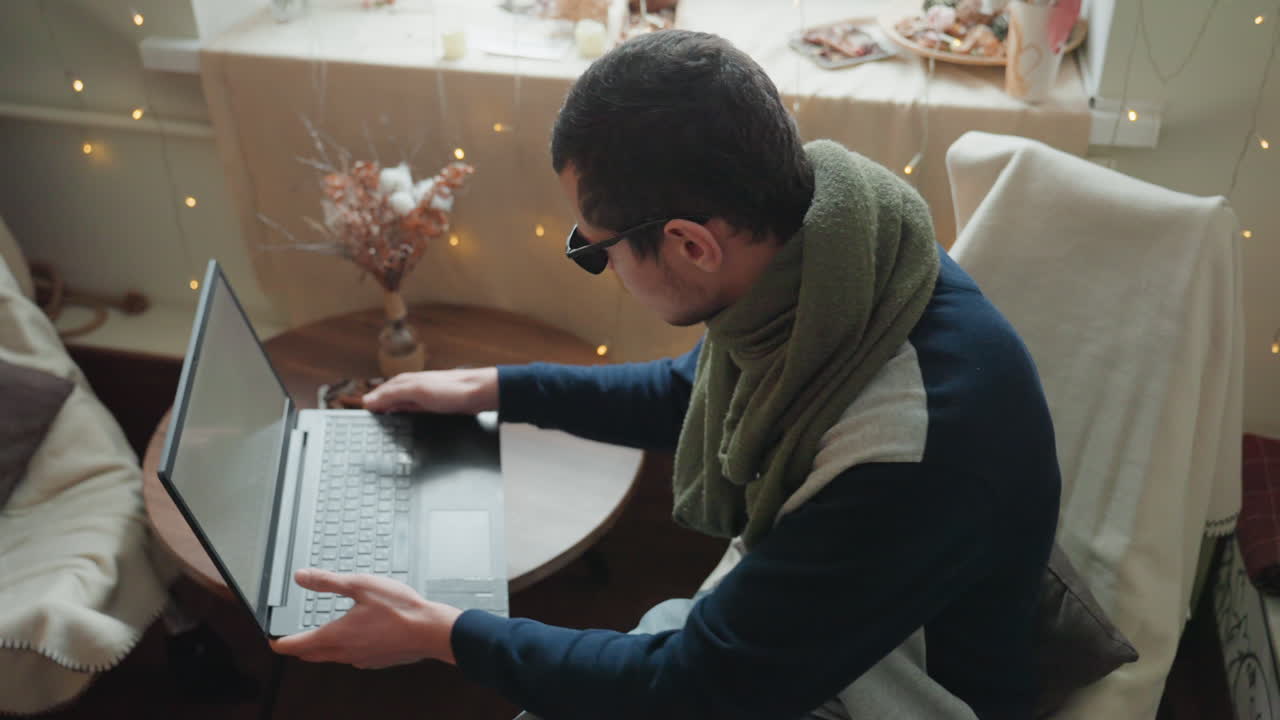 Caucasian man working on laptop at round table wearing sunglasses and green scarf, adjusting screen and typing, focused remote work vibe, fairy lights and dried flowers in background, home office