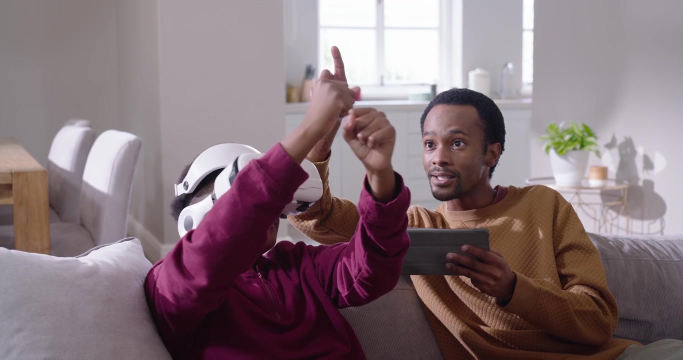 Afroamerikansk far rækker fjernbetjening og tapper på tablet, mens han guider teenagesøn iført VR-headset på sofaen