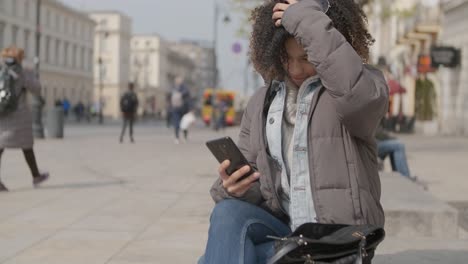 Schönes-Mädchen-Mit-Afro-Haarschnitt,-Das-Auf-Einer-Bank-An-Der-Stadtstraße-Sitzt